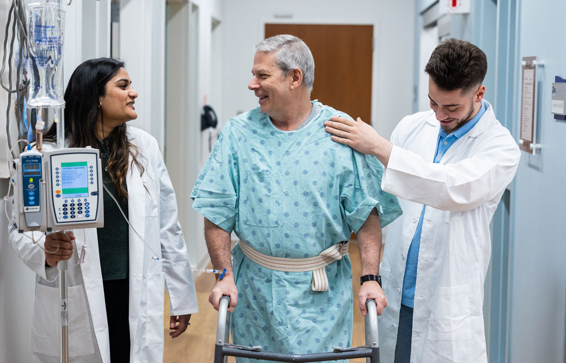 a simulation patient smiles while two students help him use a walker